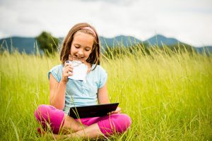 Girl Sitting In Field Enjoying A Snack While Browsing On Her Tablet